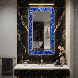 May include: A rectangular wall mirror featuring a vibrant blue, crystalline-textured frame. The mirror is mounted above a white bathroom vanity with a gold faucet, positioned against a black marble wall with gold veining and integrated lighting.