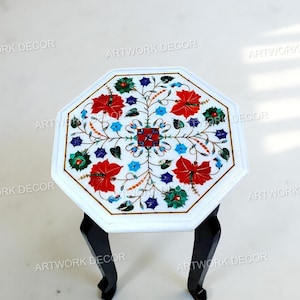 Puede incluir: Mesa octogonal de mármol blanco con un diseño floral incrustado con detalles en rojo, azul y verde. La mesa descansa sobre patas negras anguladas, mostrando una mezcla de elementos de diseño clásicos y contemporáneos. El diámetro de la mesa es de unos 61 cm.