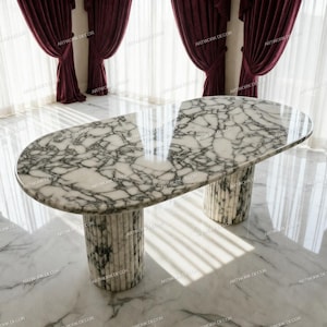 Op de afbeelding: Een ovale eettafel met een wit en grijs marmeren blad met opvallende aders. De tafel wordt ondersteund door twee cilindrische zuilen, elk opgebouwd uit verticale stenen latten die passen bij het marmerpatroon van het tafelblad.