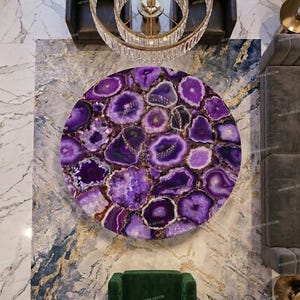 Può includere: Un tavolino da caffè circolare con un piano composto da numerose fette di agata viola lucidata con bande concentriche visibili e centri cristallini, disposte a mosaico con un bordo metallico color oro.