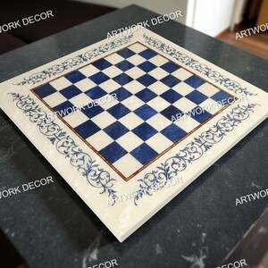 May include: Square marble chess board with a blue and white checkered pattern. The board is framed with a decorative border featuring blue floral scrollwork. The board is set on a dark surface.