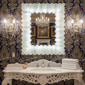 May include: A square, illuminated mirror with a white, textured frame hangs above a marble vanity. The vanity has a decorative, carved design and a built-in sink. A stack of white towels and a perfume bottle are on the vanity.
