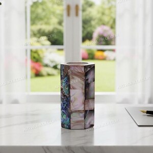 May include: A decorative, hexagonal vase with a mosaic pattern of iridescent abalone shell pieces. The vase is a mix of pink, blue, and white hues. The vase is on a white surface near a window.