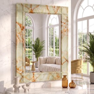 May include: A rectangular wall mirror featuring a frame with a polished light green and orange marble-like pattern. The mirror is mounted on a white marble wall above a matching white marble shelf, reflecting a room with arched windows and furniture.