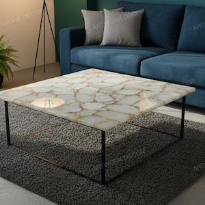 May include: A square coffee table with a white and gold patterned top and black metal legs. The table is set on a grey rug in front of a blue sofa. A potted plant is visible on the left.