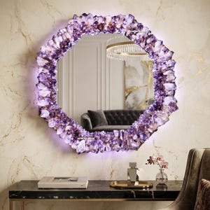 May include: A round wall mirror featuring a thick frame composed of numerous raw, purple amethyst crystal clusters. The mirror includes integrated LED backlighting that emits a soft glow around the perimeter. The text ARTWORK DECOR is visible.