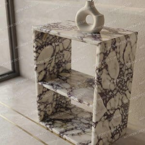 May include: A rectangular two-tier side table featuring a white and purple marble pattern with a smooth finish. A small, off-white, circular ceramic vase with a central cutout and a thin, bare, dark-coloured branch is placed on the top surface.