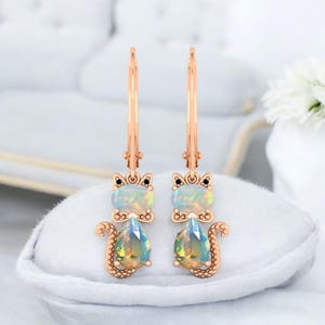 May include: A pair of rose gold drop earrings shaped like cats. Each earring features a round opal head with small black stone eyes and a pear-shaped opal body. The cat design includes a textured tail and a secure lever-back closure mechanism.
