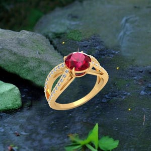 Puede incluir: Un anillo de oro con una gran gema roja redonda como pieza central. La banda se divide en dos secciones a cada lado de la gema roja, cada una con pequeñas gemas transparentes. El anillo se encuentra sobre un fondo natural borroso.