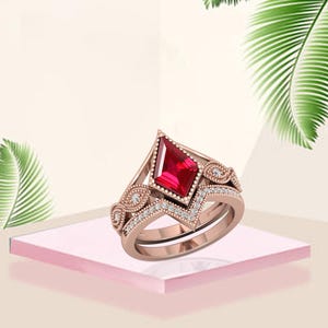 Op de afbeelding: Rosegouden ringenset met een opvallende, ruitvormige rode edelsteen. De ring is versierd met kleine, sprankelende witte stenen en decoratieve filigraan details. De set bevat een bijpassende band.