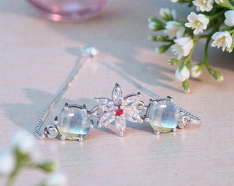 May include: A delicate silver bracelet featuring a floral centerpiece with a small red gem and clear stones. Two round, iridescent moonstone-like gems flank the flower. The bracelet is on a light-colored surface.