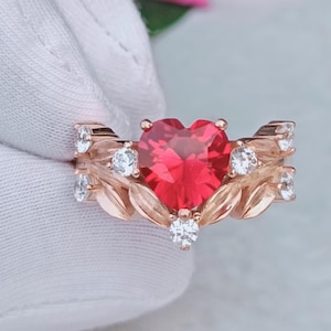 Puede incluir: Anillo de oro rosa con una piedra preciosa roja en forma de corazón. El anillo está adornado con pequeñas piedras preciosas redondas y transparentes y detalles en forma de hojas. Una joya romántica y elegante.