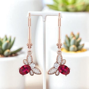 May include: Rose gold leverback earrings with a floral design. Each earring features a large oval-cut ruby gemstone, surrounded by smaller white gemstones. The earrings are displayed on a white stand against a blurred background.
