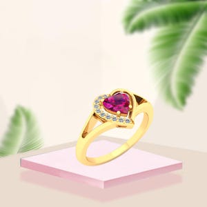 Puede incluir: Un anillo de oro con una piedra preciosa rosa oscuro en forma de corazón. El corazón está rodeado de pequeñas piedras transparentes. El anillo está sobre una superficie rosa, con un fondo de hojas verdes.
