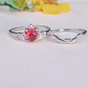 Peut inclure: Ensemble de bagues en argent avec une pierre rouge en forme de cœur et une alliance assortie. La pierre en forme de cœur est le point central, sertie dans un sertissage argenté décoratif. L'alliance a un design incurvé avec des volutes décoratives.