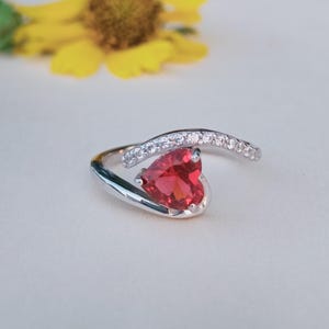 Puede incluir: Un anillo de plata con una piedra preciosa roja en forma de corazón. El anillo presenta una banda curva engastada con pequeñas piedras transparentes. El diseño es moderno y elegante, adecuado para una ocasión especial. Es una joya.