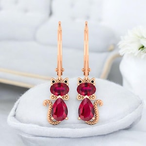 Puede incluir: Un par de pendientes en forma de gato en oro rosa. Cada pendiente presenta una gran gema roja oscura de talla ovalada para la cabeza, una gema roja en forma de pera para el cuerpo y pequeñas gemas negras para los ojos. Los pendientes tienen cierre de palanca.