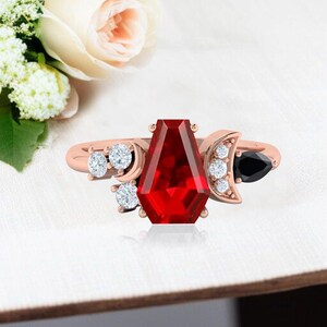 Peut inclure: Bague en or rose ornée d'une grande pierre rouge rectangulaire. La bague est ornée de pierres blanches et noires, et de détails en forme de croissant de lune. Un bijou unique et élégant.