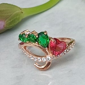 Puede incluir: Anillo de oro rosa con piedras preciosas verdes y rojas. El anillo tiene un diseño único con una piedra roja en forma de corazón y varias piedras verdes. La banda está adornada con pequeñas piedras transparentes, añadiendo brillo a la joya.