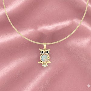 Könnte beinhalten: Goldfarbene Eulen-Anhänger-Halskette mit einem hellen Opal-Mittelstein. Die Eule hat schwarze Augen und sitzt auf einem Ast. Die Halskette ist ebenfalls goldfarben und vor einem rosa Hintergrund platziert.