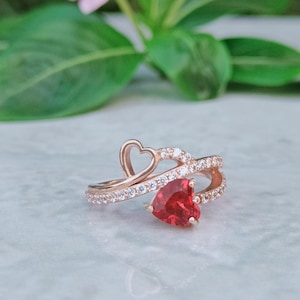 Puede incluir: Anillo de oro rosa con una piedra roja en forma de corazón y un delicado diseño de corazón abierto. La banda está adornada con pequeñas piedras claras y brillantes. Una joya romántica y elegante.