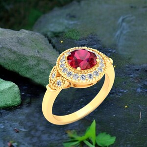 Puede incluir: Un anillo dorado con una gran gema roja redonda, rodeada de gemas claras más pequeñas. El anillo tiene un diseño detallado y ornamentado con pequeñas gemas adicionales en los lados. El fondo es borroso, con rocas y agua.