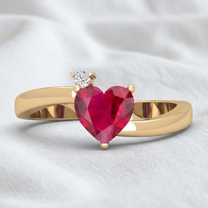 Puede incluir: Un anillo de oro con una piedra preciosa roja en forma de corazón en el centro, acentuado por un pequeño diamante blanco redondo colocado justo encima del corazón, con una banda de oro pulido y lisa que se curva alrededor de las piedras.