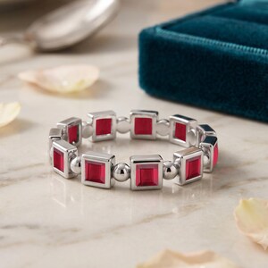 Puede incluir: Un anillo de tono plateado que presenta una serie de piedras preciosas rojas de corte cuadrado alternadas con esferas de plata pulida. La banda está construida con un patrón repetitivo de estos engastes cuadrados y cuentas metálicas redondas.