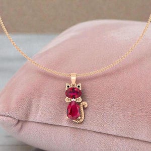 Puede incluir: Un collar con colgante de gato dorado con cuerpo y cabeza de piedra preciosa roja. El colgante de gato cuelga de una delicada cadena dorada, sobre un fondo rosa. El gato tiene ojos negros y una pajarita.