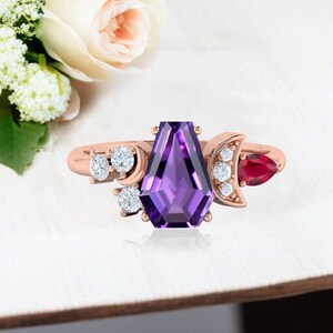 Peut inclure: Bague en or rose ornée d'une grande améthyste violette géométrique. La bague est ornée de petits diamants et d'un rubis rouge en forme de poire. Le design comprend des accents de croissant de lune.