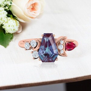 Puede incluir: Anillo de oro rosa con una gran piedra de alexandrita rectangular que cambia de color. Acentuado con diamantes y un rubí en forma de pera, el anillo tiene un diseño celestial con detalles de luna creciente y estrella. El anillo está sobre una superficie blanca.