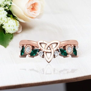 Può includere: Anello in oro rosa con design a nodo celtico, gemme verde smeraldo e bianche. L'anello presenta un nodo centrale, smeraldi a forma di marquise e gemme bianche rotonde. L'anello è posto su uno sfondo bianco morbido.