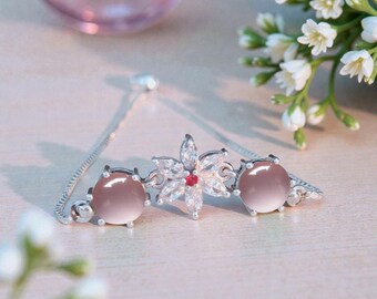 May include: A silver bracelet featuring a floral design with a red center stone and clear crystals. The bracelet has two round, pink-toned stones on either side of the flower. The bracelet is on a light-colored surface.