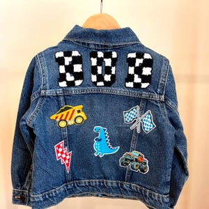 May include: A blue denim jacket with the word "DAD" in black and white checkered patches on the back. The jacket has various patches, including a yellow dump truck, a blue dinosaur, and checkered flags.