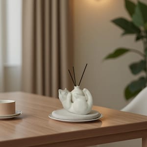May include: A white ceramic cat-shaped diffuser with a small vase holding black reeds. The diffuser sits on a white plate atop a wooden table. A cup and saucer are visible in the background, suggesting a home decor theme.