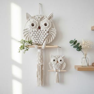 Könnte beinhalten: Zwei Wandbehänge in Form von Eulen aus Makramee in Off-White. Die größere Eule sitzt auf einer Holzstange mit Quaste. Die kleinere Eule hängt darunter. Beide haben braune Augen und befinden sich vor einer weißen Wand. Grünzeug ist auf der Stange platziert.