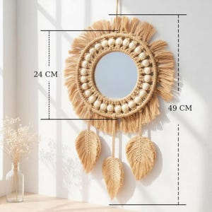 May include: A round, beige macrame mirror, 19 inches in height and 9.5 inches in width, with a central mirror and decorative fringe. Three beige leaf-shaped macrame accents hang below the mirror, adding a bohemian touch to the wall decor.