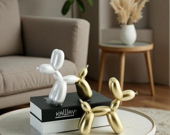 May include: Three balloon dog sculptures in white and gold tones. One white sculpture and two gold sculptures are displayed on a black and white box with the text "KAWAY!". The sculptures are on a round, light-colored table.