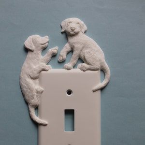 May include: White light switch cover with two white ceramic dog figurines on top. The dogs are facing each other and appear to be playing.