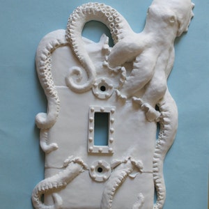 May include: White ceramic light switch cover with an octopus design. The octopus is sculpted in a 3D design with tentacles wrapping around the switch plate.