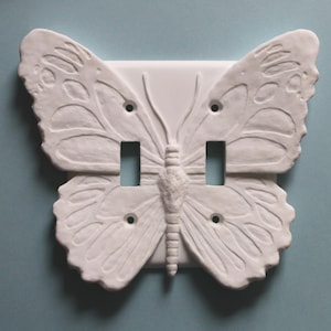 May include: White butterfly-shaped light switch cover with two switch openings. The butterfly design is raised and detailed, with visible wing patterns and a textured body. The cover is mounted on a light blue wall.