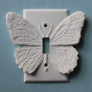 May include: A white, decorative light switch cover featuring a sculpted butterfly design. The butterfly wings are detailed with textured patterns, and the switch opening is rectangular. The cover is mounted on a light blue background.