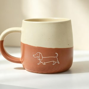 May include: A two-toned ceramic mug featuring a speckled cream upper section and a solid terracotta lower section. A simple white line drawing of a dachshund is centred on the terracotta portion, and the handle is finished in matching terracotta.