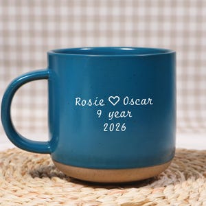 Può includere: Una tazza in ceramica color verde acqua con finitura maculata e base color sabbia naturale. Il lato anteriore presenta una scritta bianca che riporta Rosie, un simbolo di cuore, Oscar, 9 year e 2026. La tazza ha un manico curvo a sinistra.