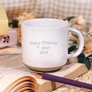 Op de afbeelding: Witte keramische mok met een bruine basis, gegraveerd met "Grace ♡ Harry 9 year 2025." Een paarse pen ligt op een open boek. De achtergrond bevat bloemen en een neutraal gekleurd patroon.