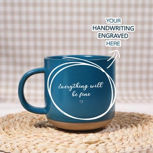 Puede incluir: Una taza de cerámica color turquesa con base marrón, con el texto "Everything will be fine" en escritura blanca, con un pequeño símbolo de corazón. La taza está sobre un posavasos tejido. La parte superior de la taza tiene el texto "YOUR HANDWRITING ENGRAVED HERE".