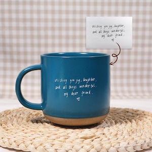 Puede incluir: Una taza de cerámica color turquesa con base marrón y asa curva. La taza tiene el texto escrito a mano "Wishing you joy, laughter, and all things wonderful, my dear friend." Se adjunta una pequeña nota con el mismo texto.