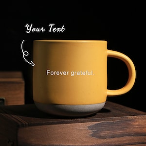 Puede incluir: Una taza de cerámica amarilla con una base gris y un asa curva. La taza tiene el texto "Forever grateful." impreso en la parte delantera. Las palabras "Your Text" están escritas encima de la taza.