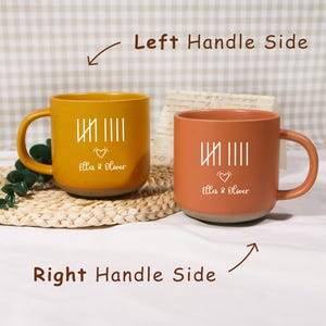 May include: Two ceramic mugs, one mustard yellow and one terracotta orange, with a white design and the text "Ella & Oliver." The mugs have a rounded shape, a handle, and a speckled base. The text is accompanied by a heart symbol.