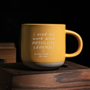 Op de afbeelding: Een gele keramische mok met een grijze basis en een gebogen handvat. De mok heeft witte tekst "i used to work with ABSOLUTE LEGENDS" met een ster en de tekst "yes, they bought me this mug..."